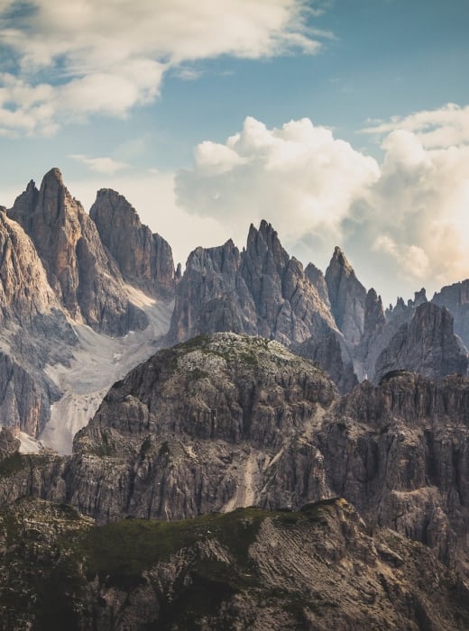 Jagged mountain peaks rise dramatically above rocky, green lower hills under a vast, cloudy sky, revealing distant valleys.