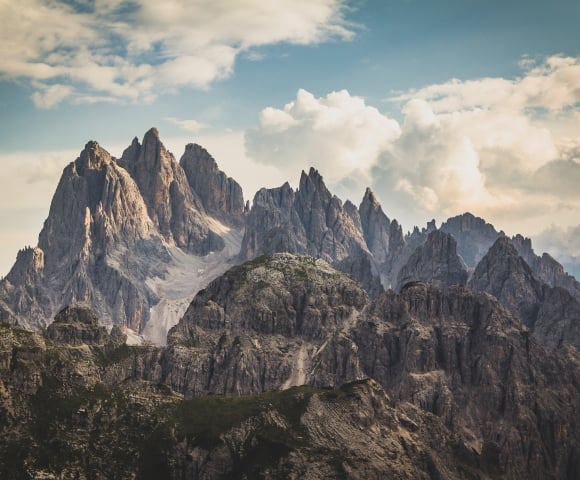 Jagged mountain peaks rise dramatically above rocky, green lower hills under a vast, cloudy sky, revealing distant valleys.