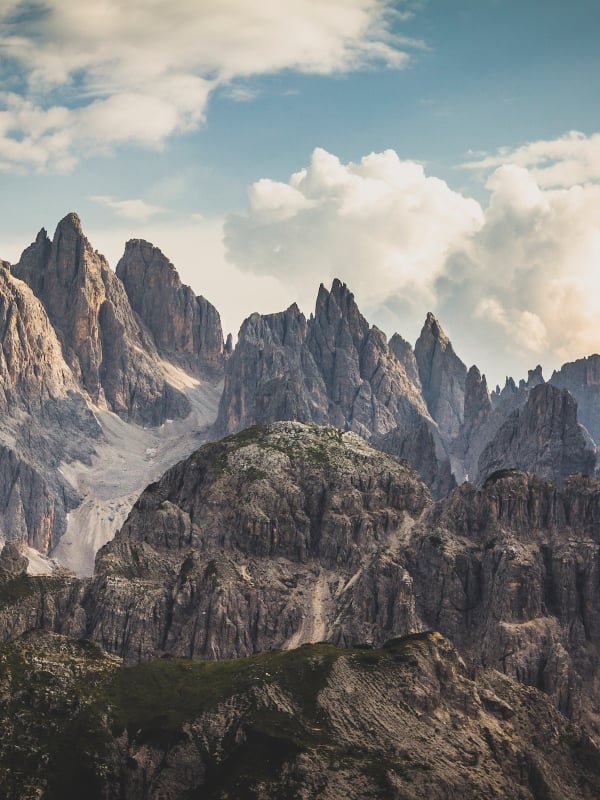 Jagged mountain peaks rise dramatically above rocky, green lower hills under a vast, cloudy sky, revealing distant valleys.