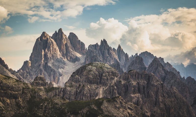 Jagged mountain peaks rise dramatically above rocky, green lower hills under a vast, cloudy sky, revealing distant valleys.