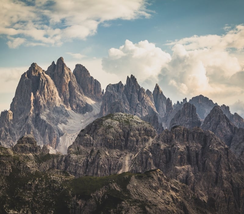 Jagged mountain peaks rise dramatically above rocky, green lower hills under a vast, cloudy sky, revealing distant valleys.