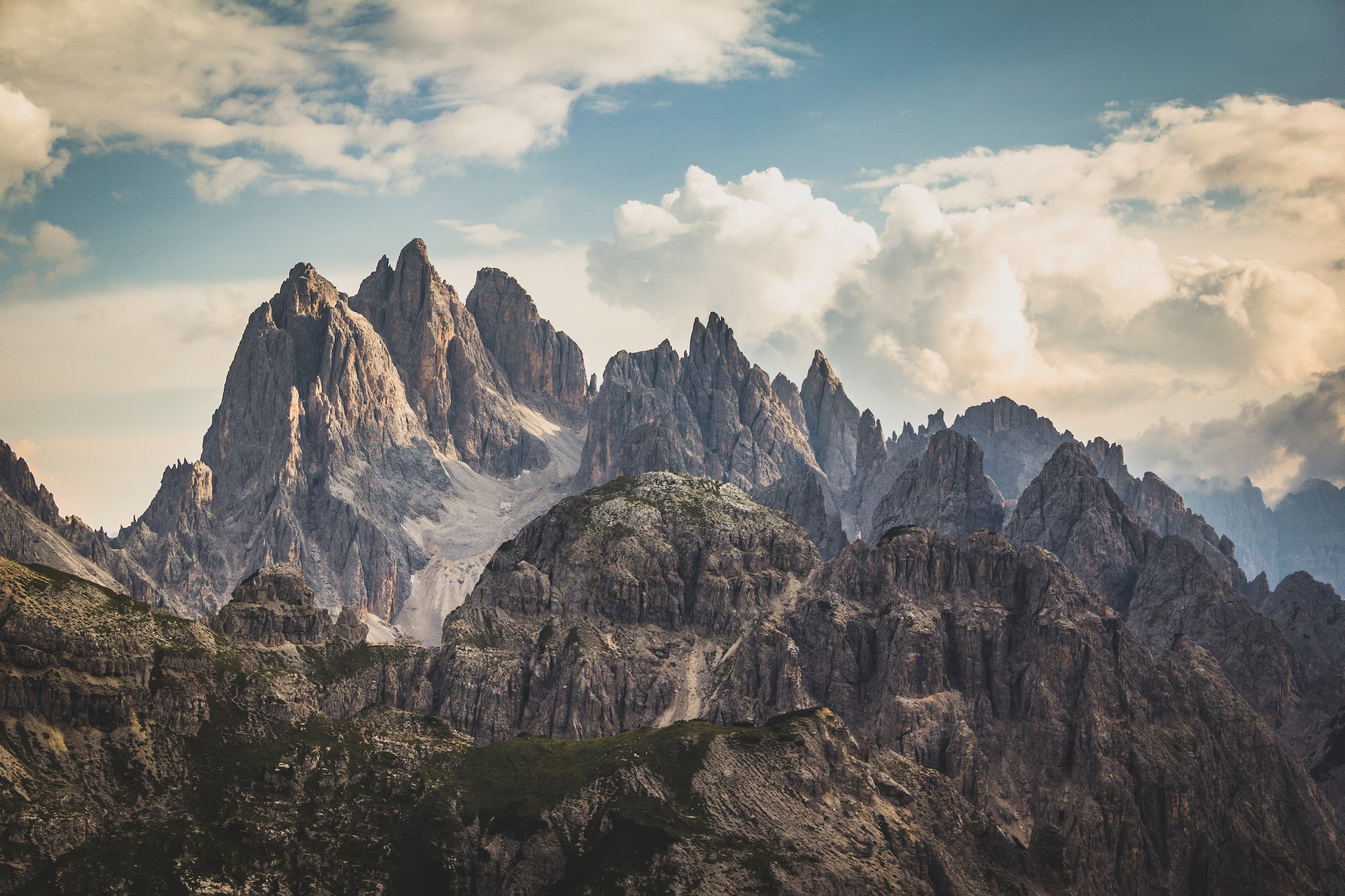 Jagged mountain peaks rise dramatically above rocky, green lower hills under a vast, cloudy sky, revealing distant valleys.