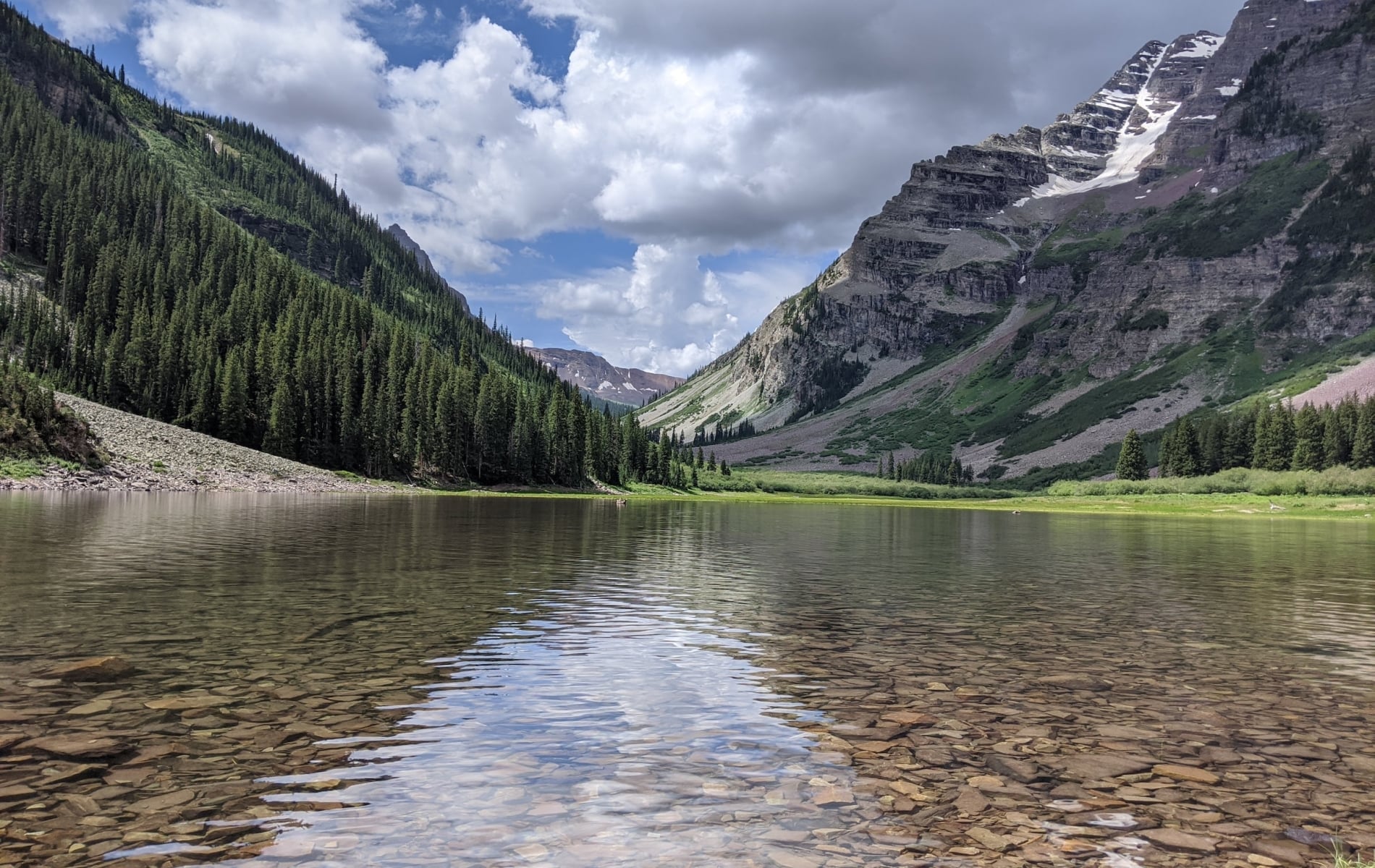 A clear mountain lake with a visible rocky bottom reflects a partly cloudy sky, nestled between two towering, forested mountains with snow patches.