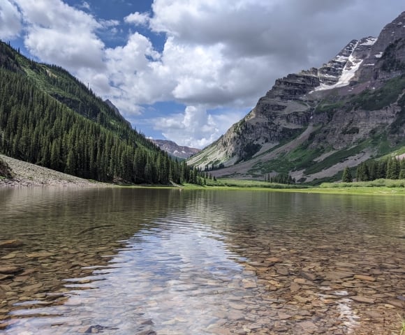 A clear mountain lake with a visible rocky bottom reflects a partly cloudy sky, nestled between two towering, forested mountains with snow patches.