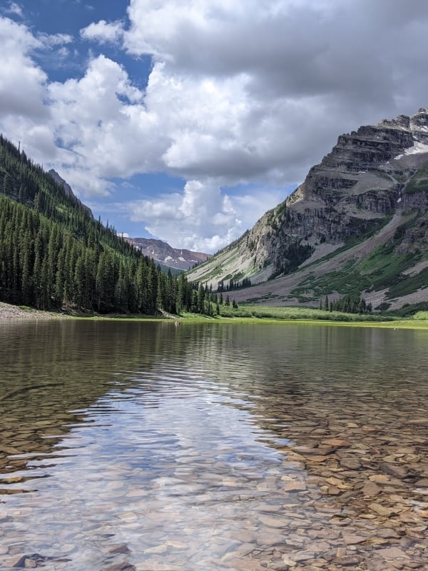 A clear mountain lake with a visible rocky bottom reflects a partly cloudy sky, nestled between two towering, forested mountains with snow patches.