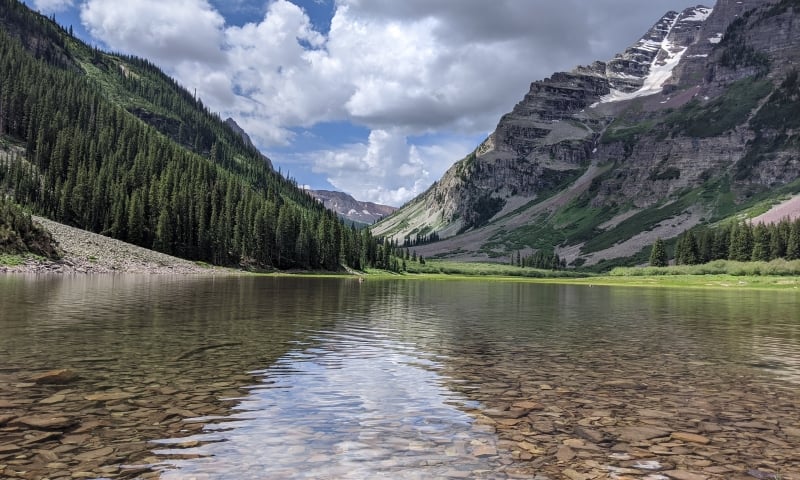 A clear mountain lake with a visible rocky bottom reflects a partly cloudy sky, nestled between two towering, forested mountains with snow patches.