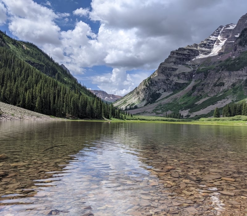A clear mountain lake with a visible rocky bottom reflects a partly cloudy sky, nestled between two towering, forested mountains with snow patches.