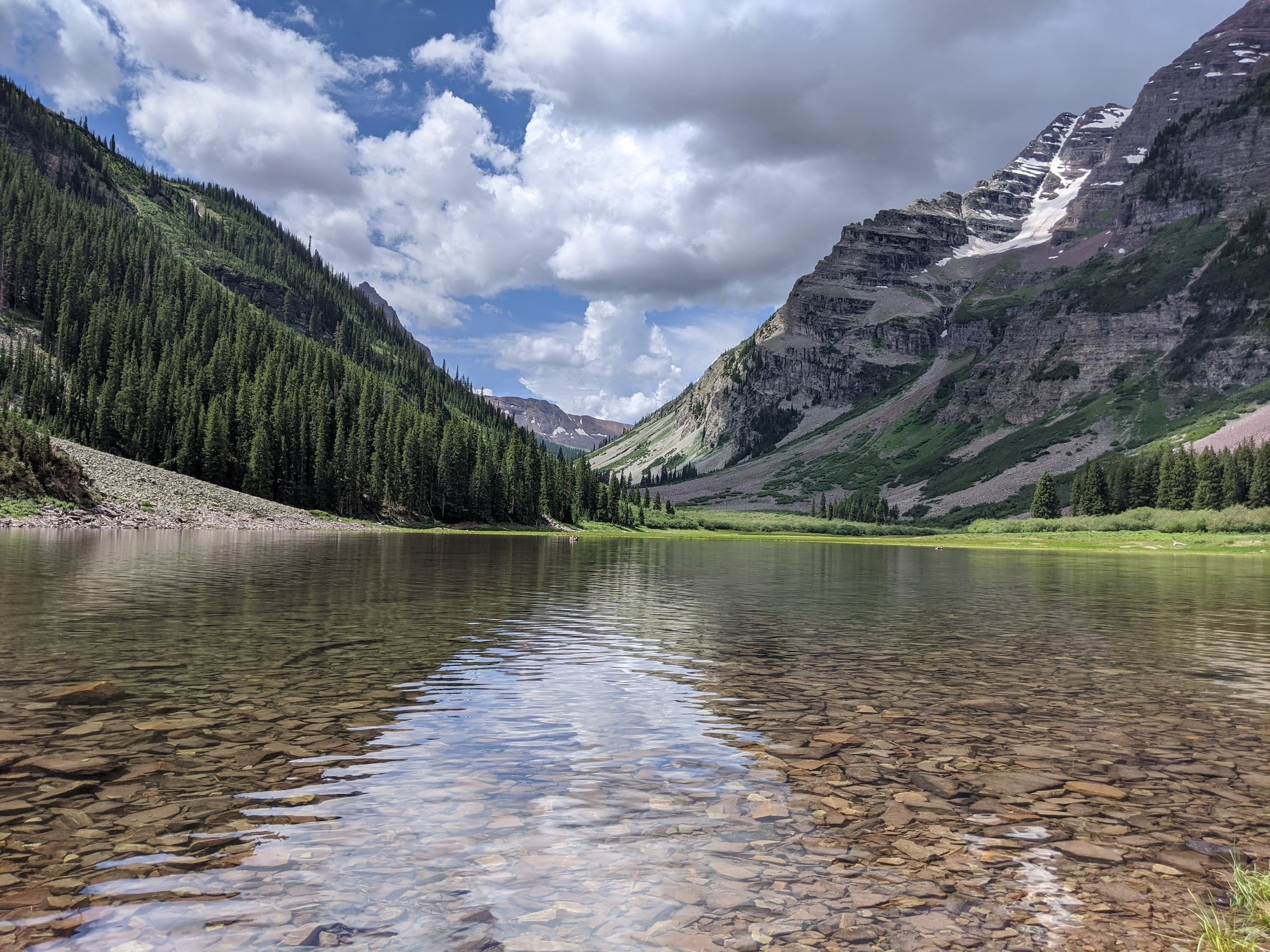 A clear mountain lake with a visible rocky bottom reflects a partly cloudy sky, nestled between two towering, forested mountains with snow patches.