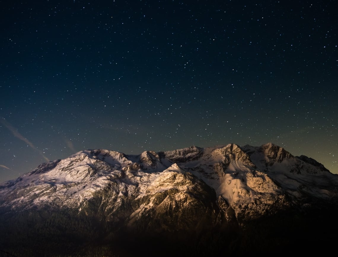 Snow-capped mountains stand illuminated, revealing textured peaks above a dark forest. Countless stars fill the deep blue night sky, with a faint glow near the horizon.