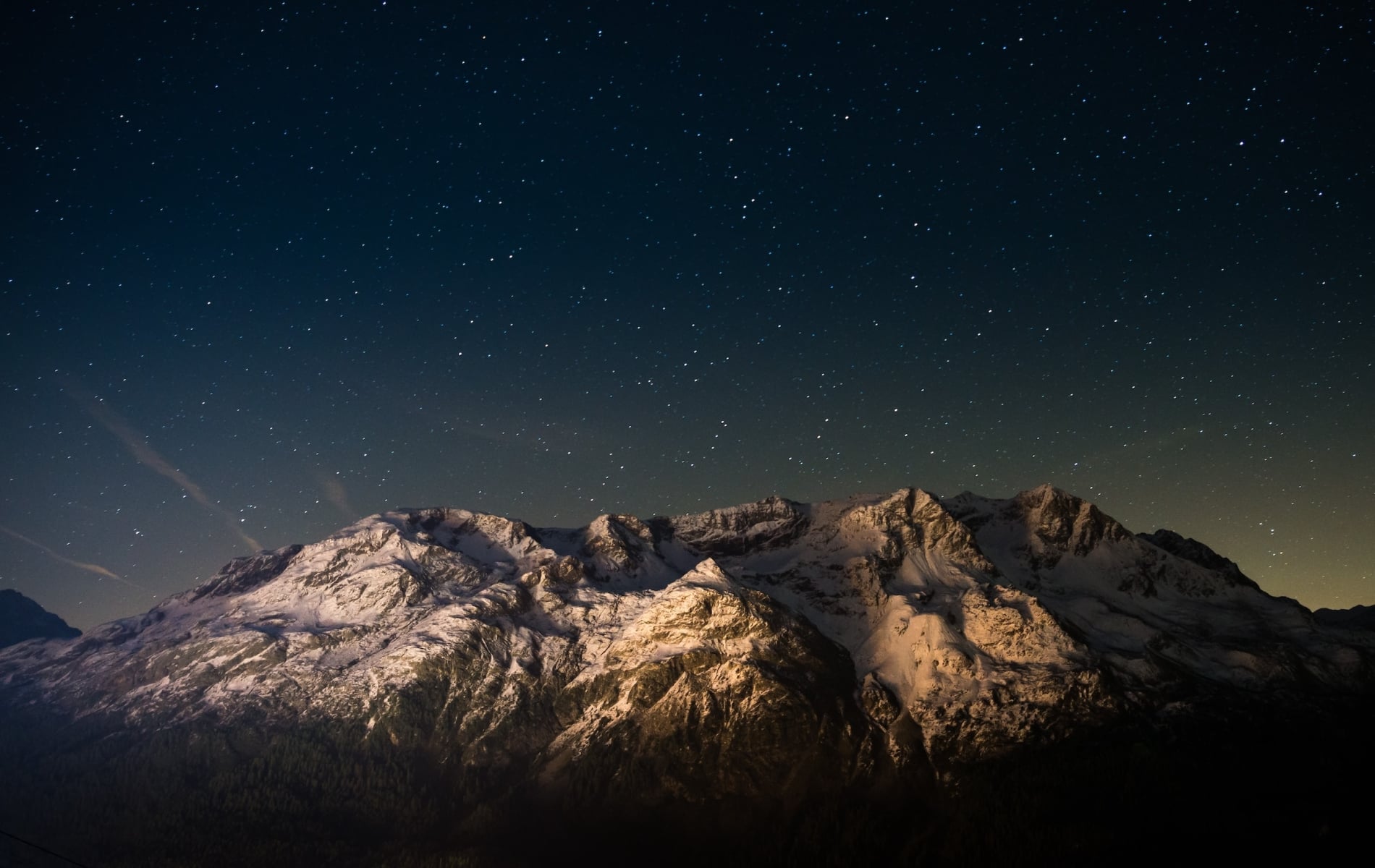 Snow-capped mountains stand illuminated, revealing textured peaks above a dark forest. Countless stars fill the deep blue night sky, with a faint glow near the horizon.