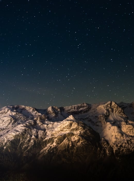 Snow-capped mountains stand illuminated, revealing textured peaks above a dark forest. Countless stars fill the deep blue night sky, with a faint glow near the horizon.