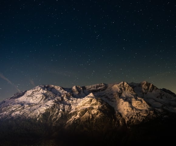 Snow-capped mountains stand illuminated, revealing textured peaks above a dark forest. Countless stars fill the deep blue night sky, with a faint glow near the horizon.