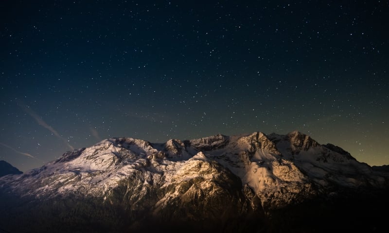 Snow-capped mountains stand illuminated, revealing textured peaks above a dark forest. Countless stars fill the deep blue night sky, with a faint glow near the horizon.