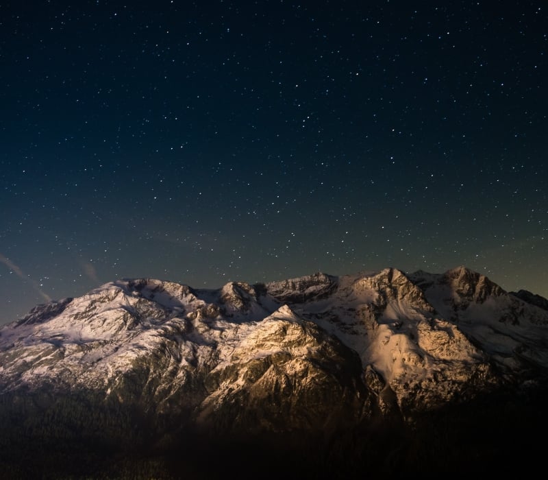 Snow-capped mountains stand illuminated, revealing textured peaks above a dark forest. Countless stars fill the deep blue night sky, with a faint glow near the horizon.