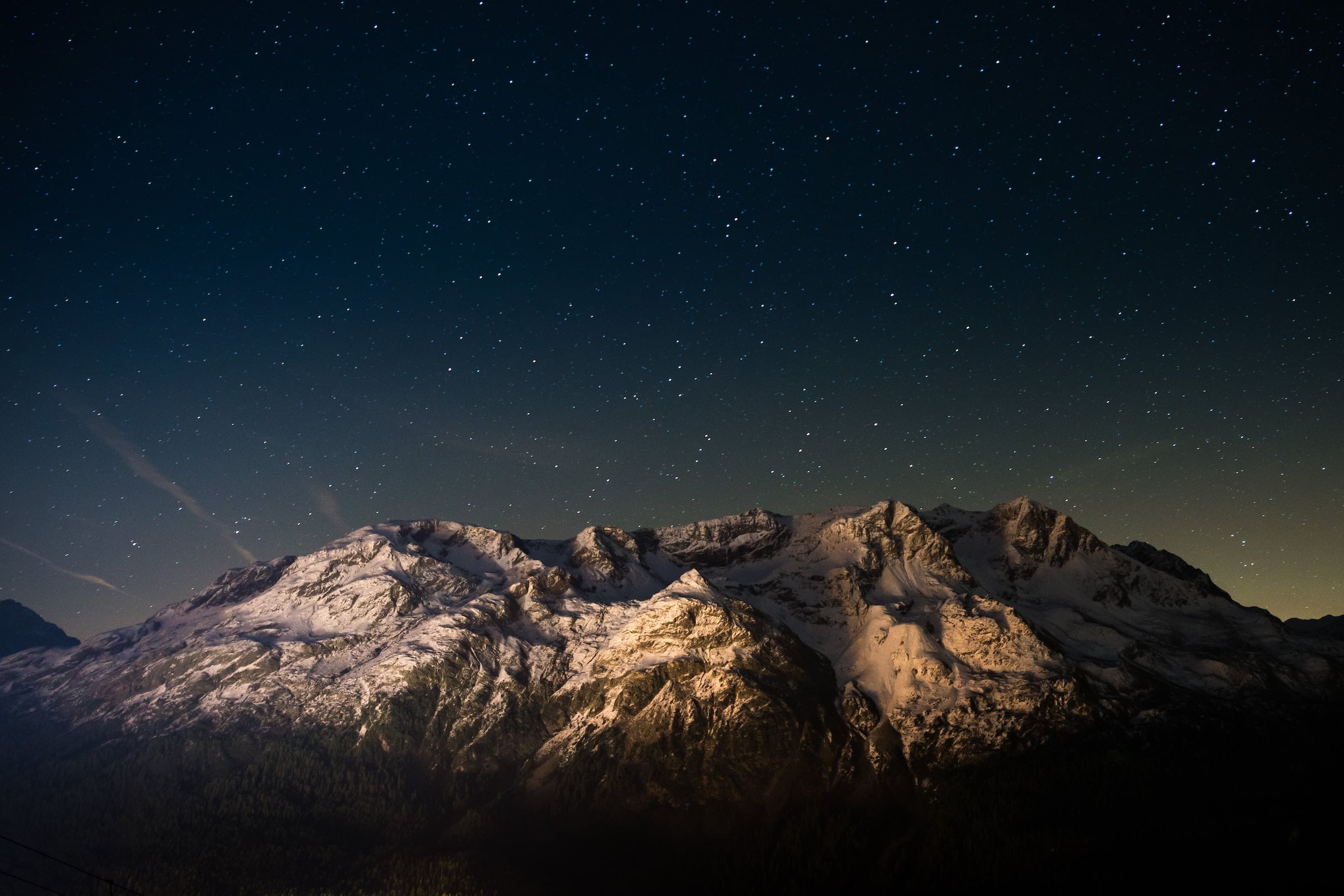 Snow-capped mountains stand illuminated, revealing textured peaks above a dark forest. Countless stars fill the deep blue night sky, with a faint glow near the horizon.