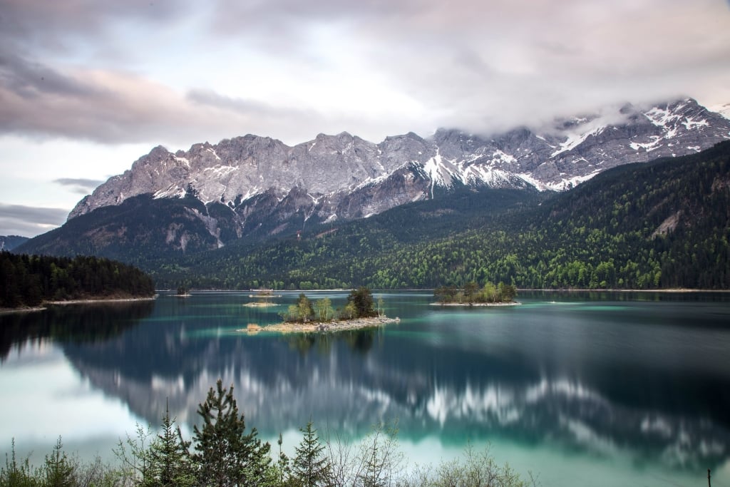Turquoise lake reflects snow-capped mountains partially obscured by clouds, with several tree-covered islands, surrounded by evergreen forests under a grey sky.