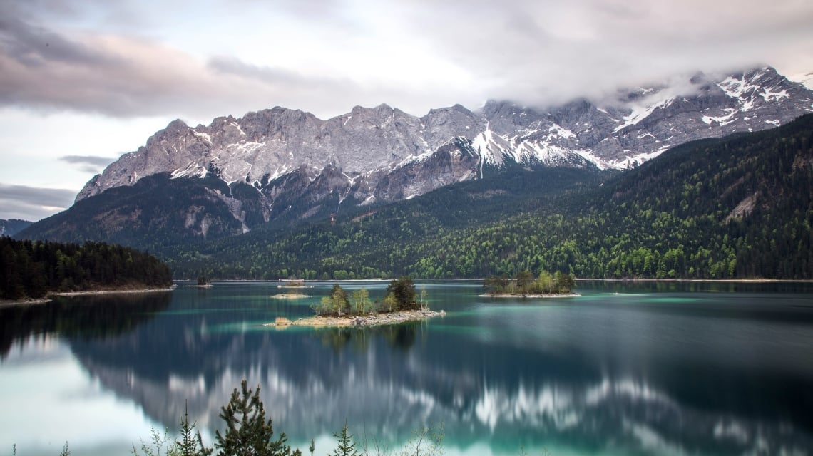 Turquoise lake reflects snow-capped mountains partially obscured by clouds, with several tree-covered islands, surrounded by evergreen forests under a grey sky.