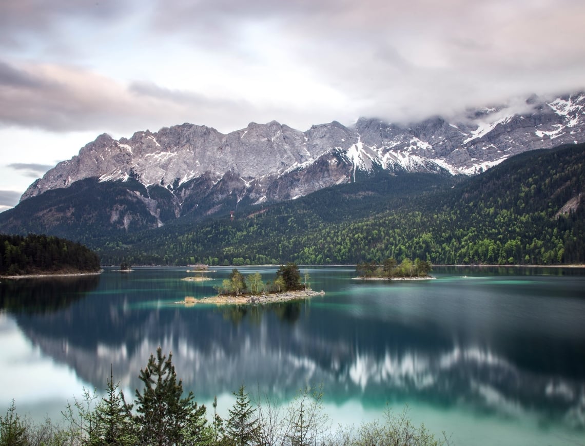Turquoise lake reflects snow-capped mountains partially obscured by clouds, with several tree-covered islands, surrounded by evergreen forests under a grey sky.