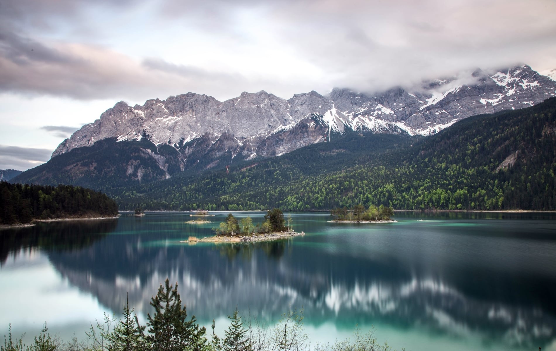 Turquoise lake reflects snow-capped mountains partially obscured by clouds, with several tree-covered islands, surrounded by evergreen forests under a grey sky.