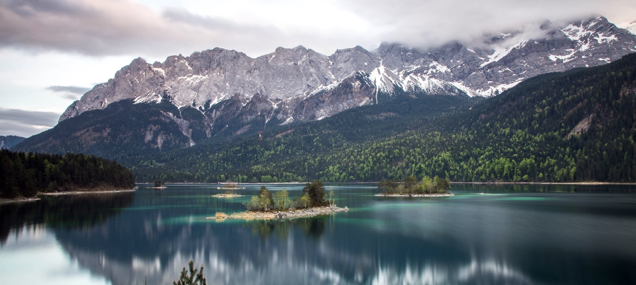 Turquoise lake reflects snow-capped mountains partially obscured by clouds, with several tree-covered islands, surrounded by evergreen forests under a grey sky.