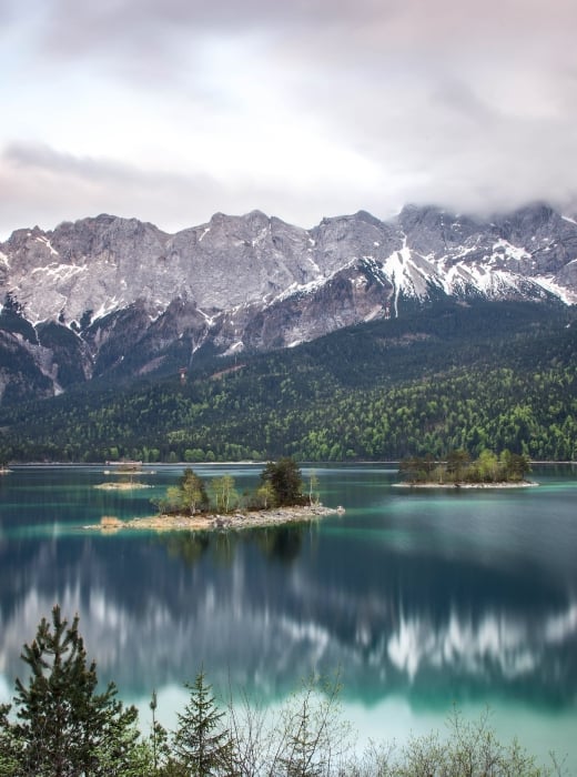Turquoise lake reflects snow-capped mountains partially obscured by clouds, with several tree-covered islands, surrounded by evergreen forests under a grey sky.