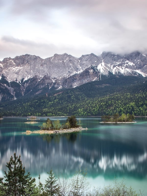 Turquoise lake reflects snow-capped mountains partially obscured by clouds, with several tree-covered islands, surrounded by evergreen forests under a grey sky.