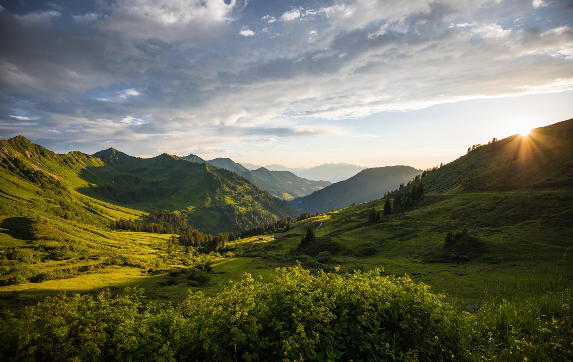 A vast mountain landscape features multiple green peaks and valleys, with the setting sun casting dramatic light from the right under a cloudy sky.
