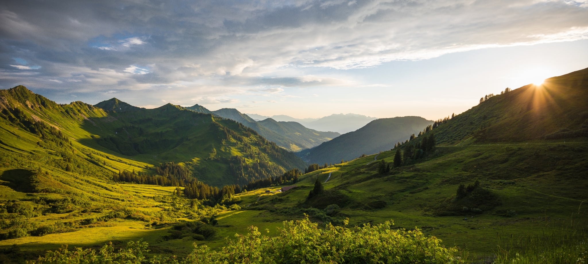 A vast mountain landscape features multiple green peaks and valleys, with the setting sun casting dramatic light from the right under a cloudy sky.