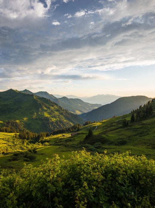 A vast mountain landscape features multiple green peaks and valleys, with the setting sun casting dramatic light from the right under a cloudy sky.