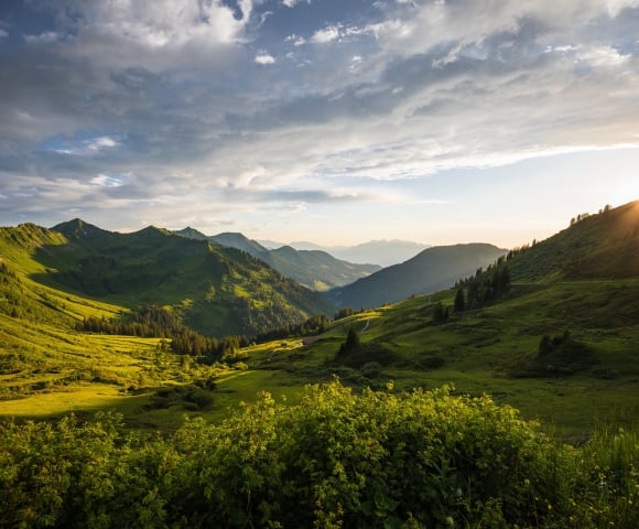 A vast mountain landscape features multiple green peaks and valleys, with the setting sun casting dramatic light from the right under a cloudy sky.