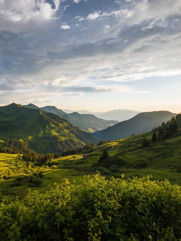 A vast mountain landscape features multiple green peaks and valleys, with the setting sun casting dramatic light from the right under a cloudy sky.