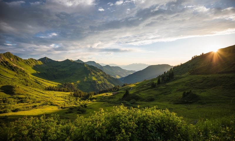 A vast mountain landscape features multiple green peaks and valleys, with the setting sun casting dramatic light from the right under a cloudy sky.