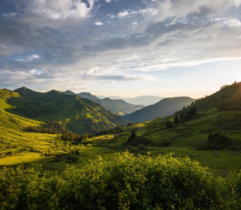 A vast mountain landscape features multiple green peaks and valleys, with the setting sun casting dramatic light from the right under a cloudy sky.