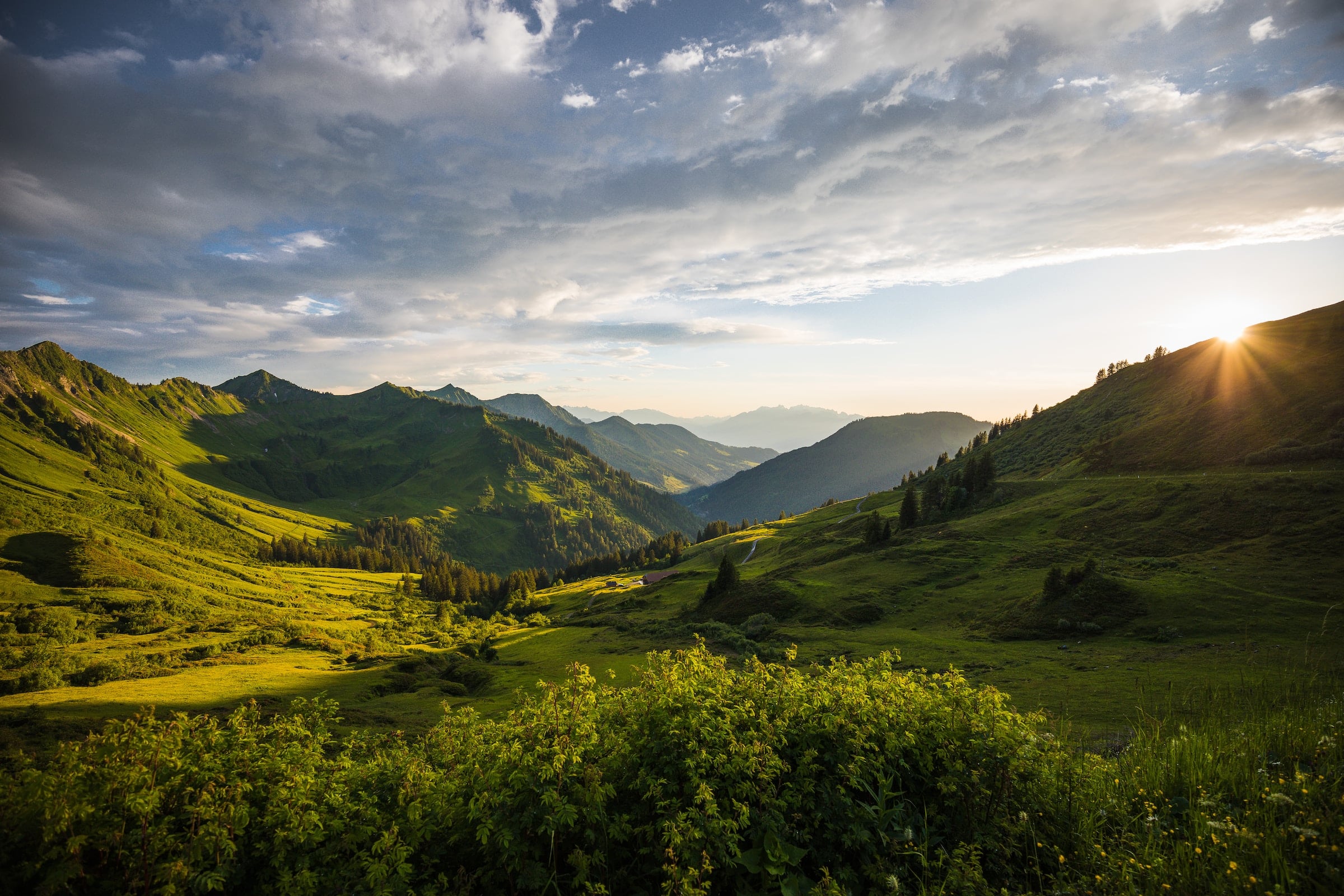 A vast mountain landscape features multiple green peaks and valleys, with the setting sun casting dramatic light from the right under a cloudy sky.
