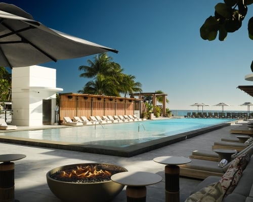 A long, inviting resort pool area features a lit fire pit, numerous lounge chairs, and beach umbrellas, all overlooking the ocean under a clear blue sky.