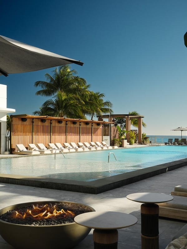 A long, inviting resort pool area features a lit fire pit, numerous lounge chairs, and beach umbrellas, all overlooking the ocean under a clear blue sky.
