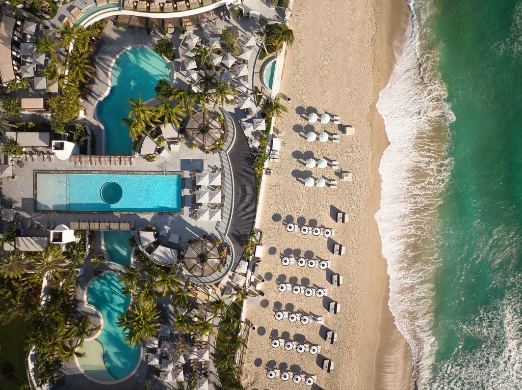 A luxury resort with multiple turquoise pools and palm trees sprawls alongside a wide sandy beach, where gentle waves meet the shore.