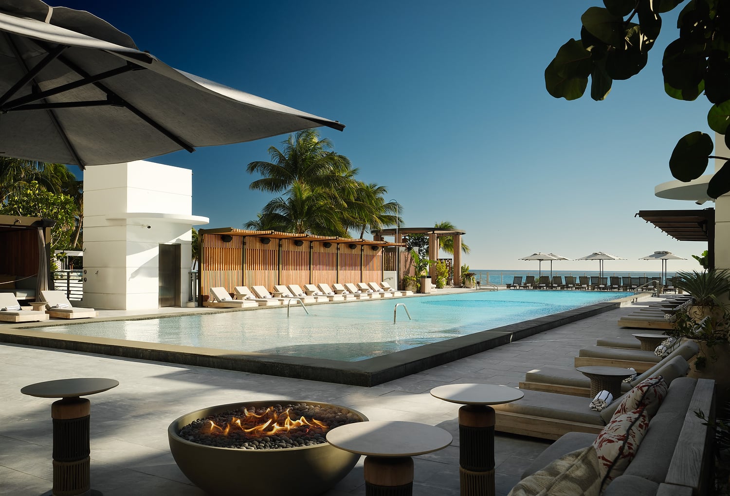 A long, inviting resort pool area features a lit fire pit, numerous lounge chairs, and beach umbrellas, all overlooking the ocean under a clear blue sky.