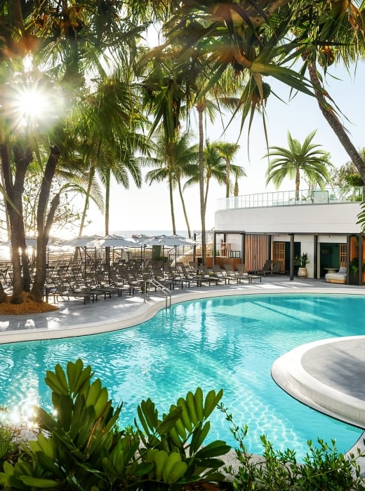 A resort pool gleams under sunshine, surrounded by numerous palm trees, lounge chairs, and a modern white building, with a distant beach and ocean visible.