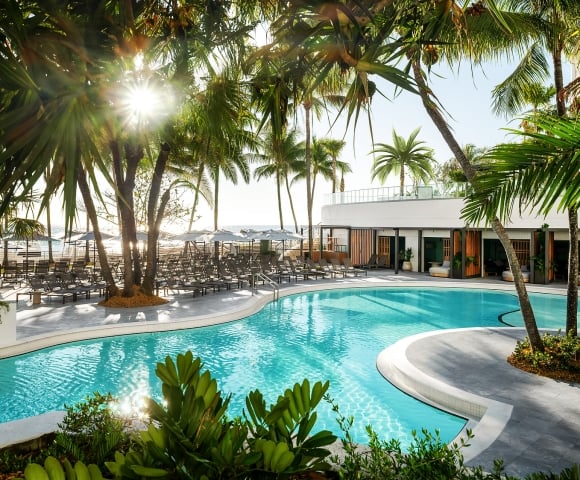 A resort pool gleams under sunshine, surrounded by numerous palm trees, lounge chairs, and a modern white building, with a distant beach and ocean visible.