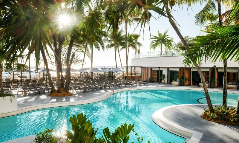 A resort pool gleams under sunshine, surrounded by numerous palm trees, lounge chairs, and a modern white building, with a distant beach and ocean visible.