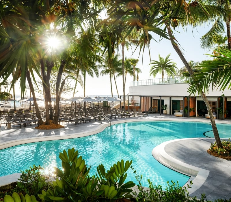 A resort pool gleams under sunshine, surrounded by numerous palm trees, lounge chairs, and a modern white building, with a distant beach and ocean visible.