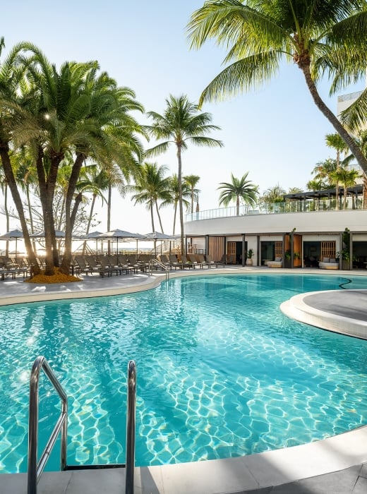 A large, winding swimming pool glistens under sunlight, surrounded by numerous palm trees, lounge chairs, and resort buildings, with the ocean in the background.