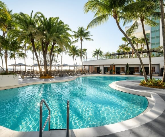 A large, winding swimming pool glistens under sunlight, surrounded by numerous palm trees, lounge chairs, and resort buildings, with the ocean in the background.