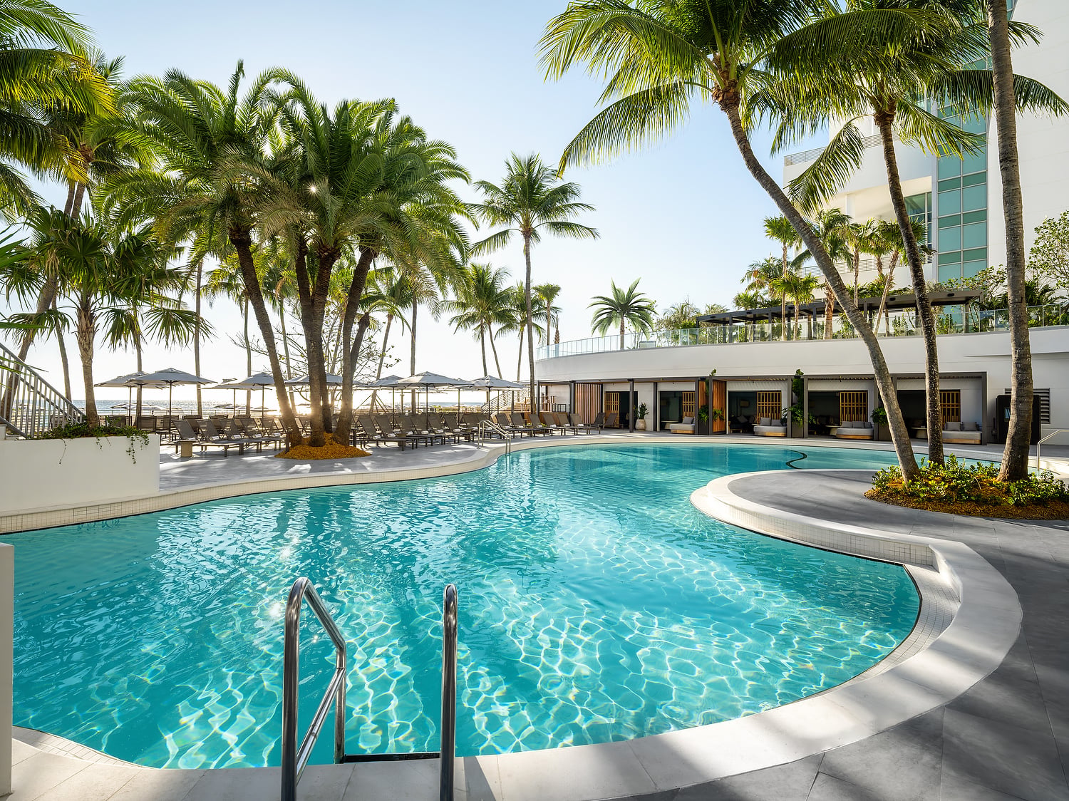 A large, winding swimming pool glistens under sunlight, surrounded by numerous palm trees, lounge chairs, and resort buildings, with the ocean in the background.