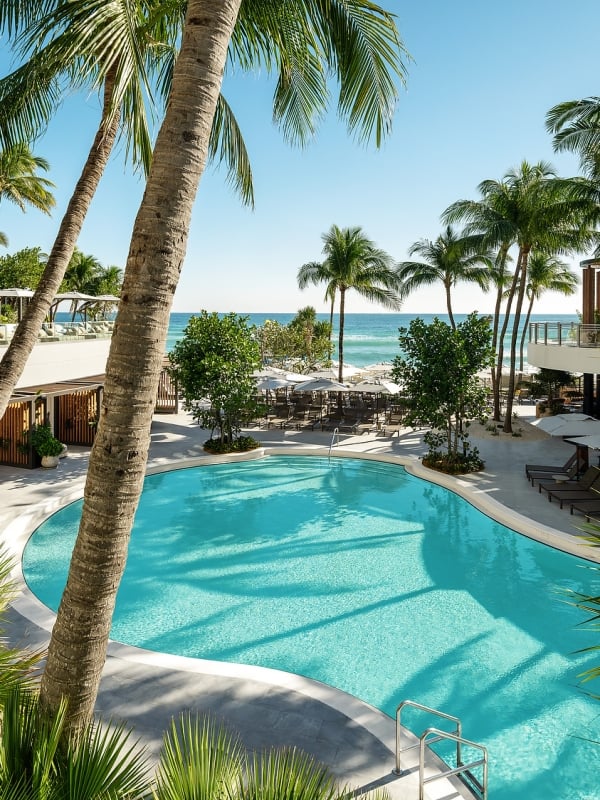 A curving blue swimming pool is surrounded by resort buildings and numerous palm trees, overlooking a sandy beach and the clear blue ocean.
