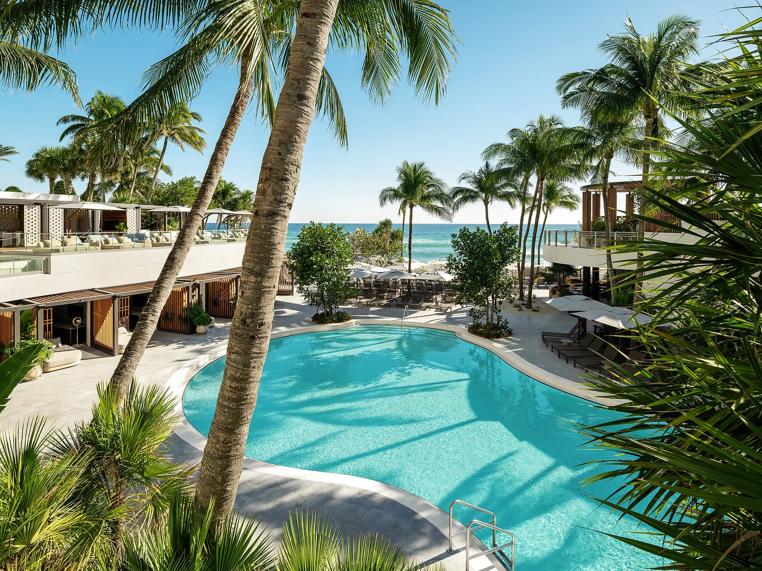 A curving blue swimming pool is surrounded by resort buildings and numerous palm trees, overlooking a sandy beach and the clear blue ocean.