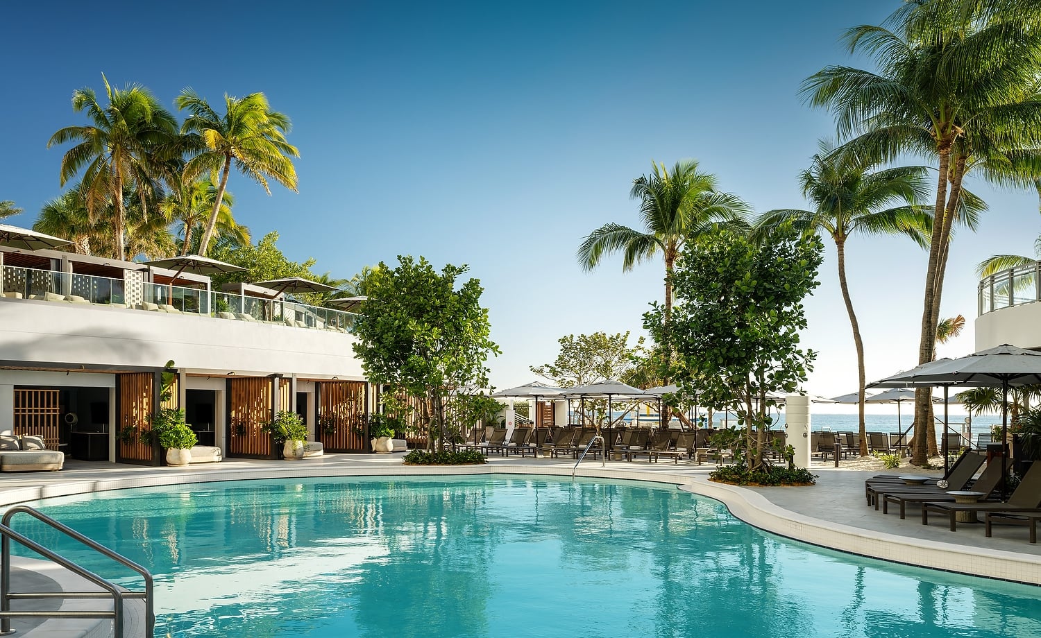 A large, turquoise swimming pool curves past a modern white resort building. Surrounded by palm trees, lounge chairs, and umbrellas, the ocean is visible under a clear blue sky.