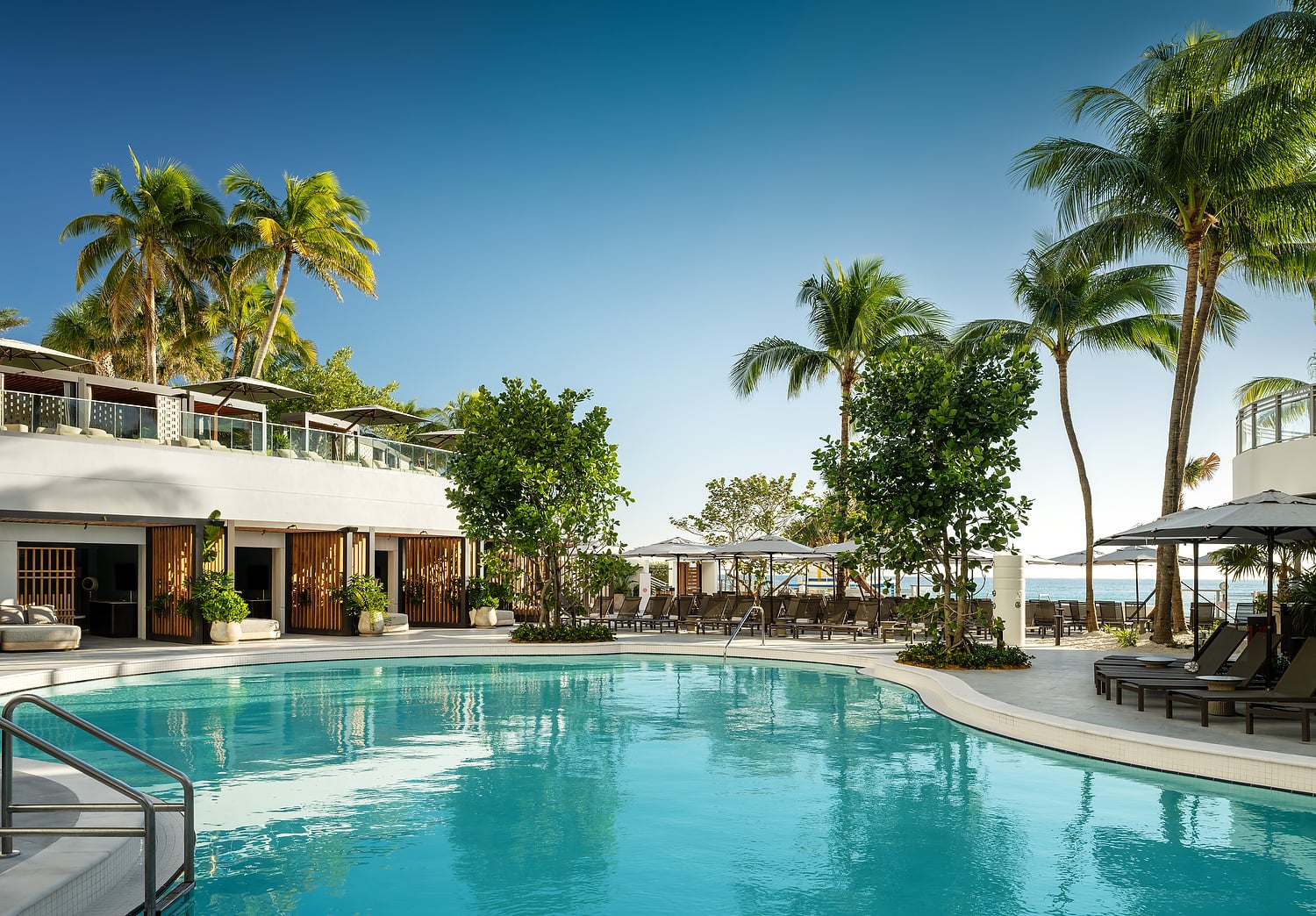 A large, turquoise swimming pool curves past a modern white resort building. Surrounded by palm trees, lounge chairs, and umbrellas, the ocean is visible under a clear blue sky.