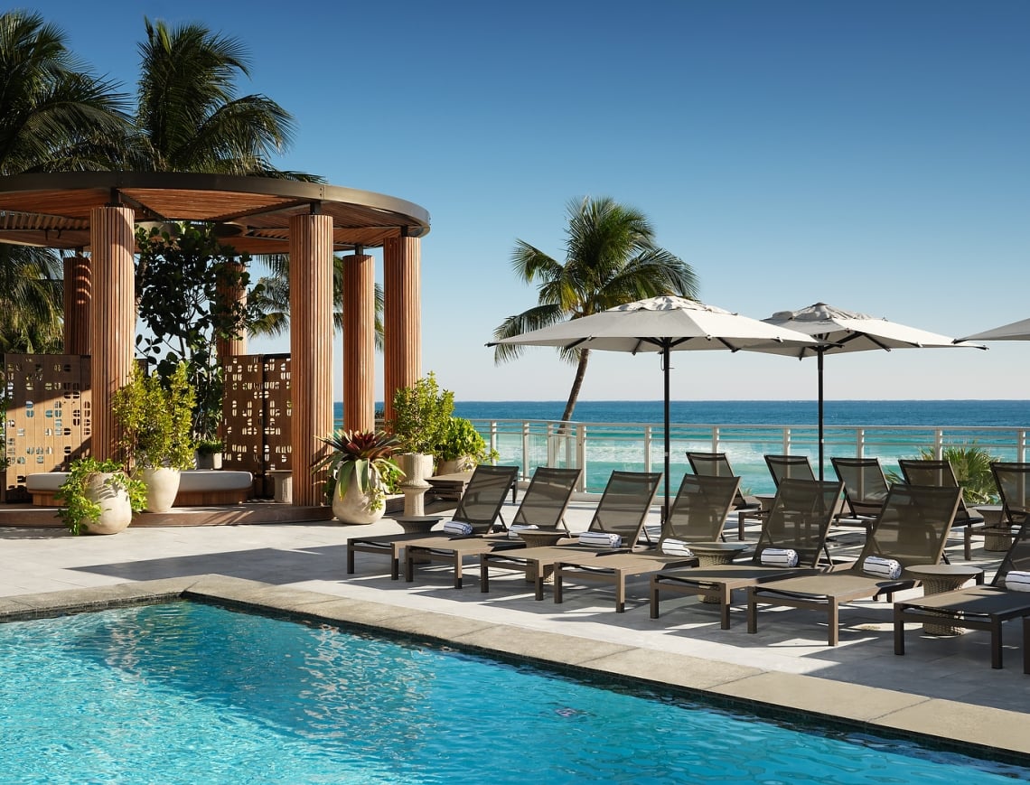 Lounge chairs with towels are arranged beside a blue swimming pool, on a sunny deck overlooking a vibrant ocean, with a wooden cabana.